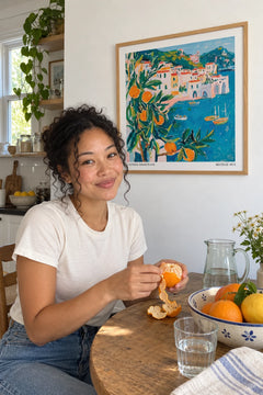 Woman peeling an orange at her kitchen table below the Costiera Amalfitana print
