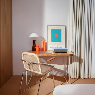 Wall office art of orange and blue squares leaning on a wooden desk with a white lamp and chair nearby