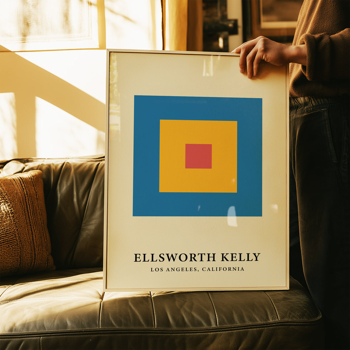 Framed minimalist art with concentric blue, yellow and red blocks leaning on a leather sofa with cushions