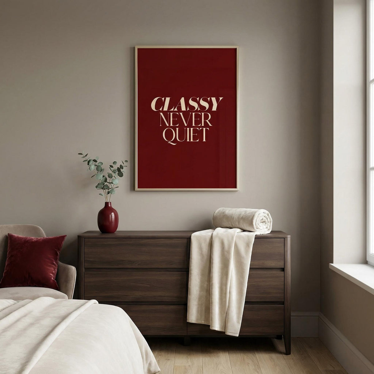 Classy wall art with beige text on dark red above a wooden dresser with a red vase and green foliage