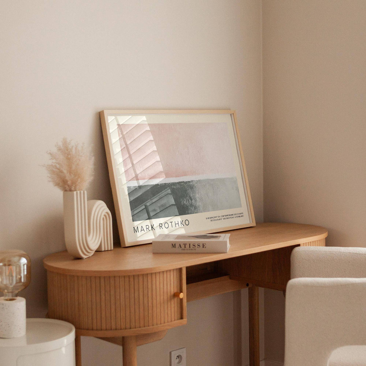 Print leaning on wooden desk of abstract blocks in muted pink and grey with a book nearby