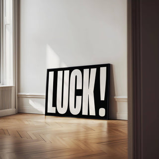 Print leaning on floor featuring the word luck in bold white letters on black background with wooden herringbone floor