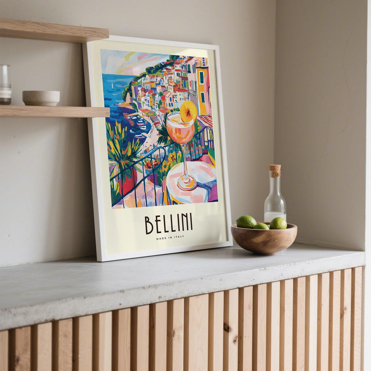 Print leaning on a shelf of a Bellini cocktail and colourful coastal village in illustration style with bowl of limes nearby