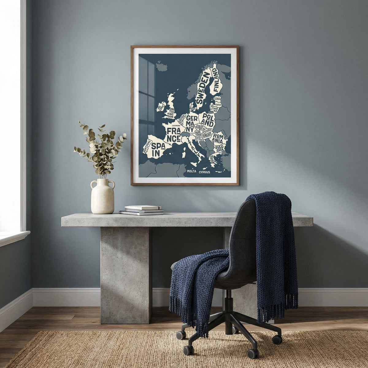 Print of Europe Map Print above a table with a white vase and books in a blue grey room