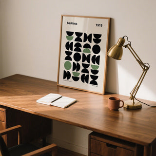 Gallery print of geometric abstract shapes in black and olive leaning on a wooden desk with a gold desk lamp