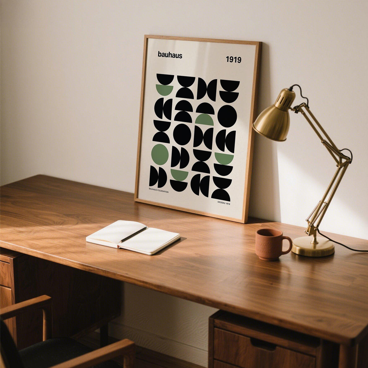Gallery print of geometric abstract shapes in black and olive leaning on a wooden desk with a gold desk lamp