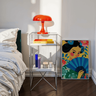 Geisha Pop Art print leaning on a wooden floor beside a metal side table with a red lamp and candles in a bedroom