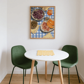 Picnic Decor Wall Art of grapes melon and olives on a blue check tablecloth in a white dining nook with green chairs