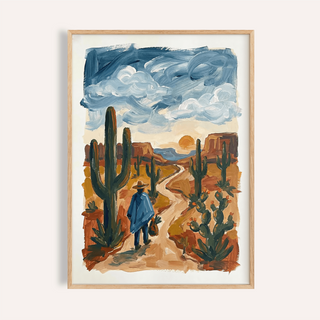 Framed print of desert wall art with a figure in blue walking down a winding path among cacti under a cloudy sky