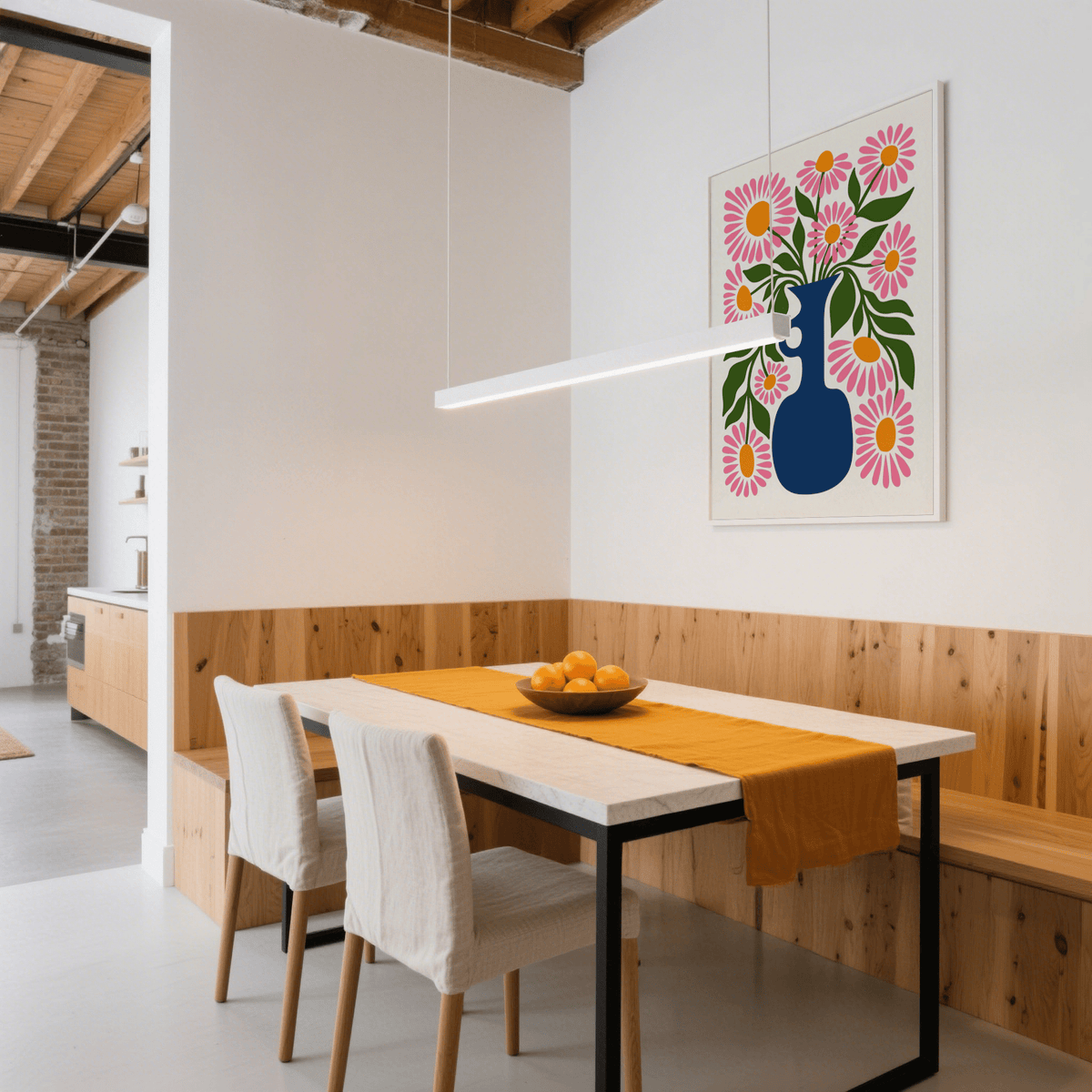 Print on a wall in a dining room with pink daisies in a blue vase above a table with a bowl of oranges