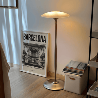 Print leaning on a floor near a minimalist metal lamp and a small white box with stacked books