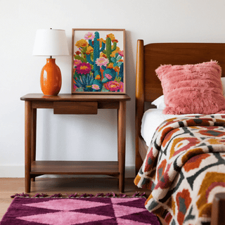 Print of cactus flower art with vibrant blue and yellow tones leaning on a wooden bedside table beside an orange lamp