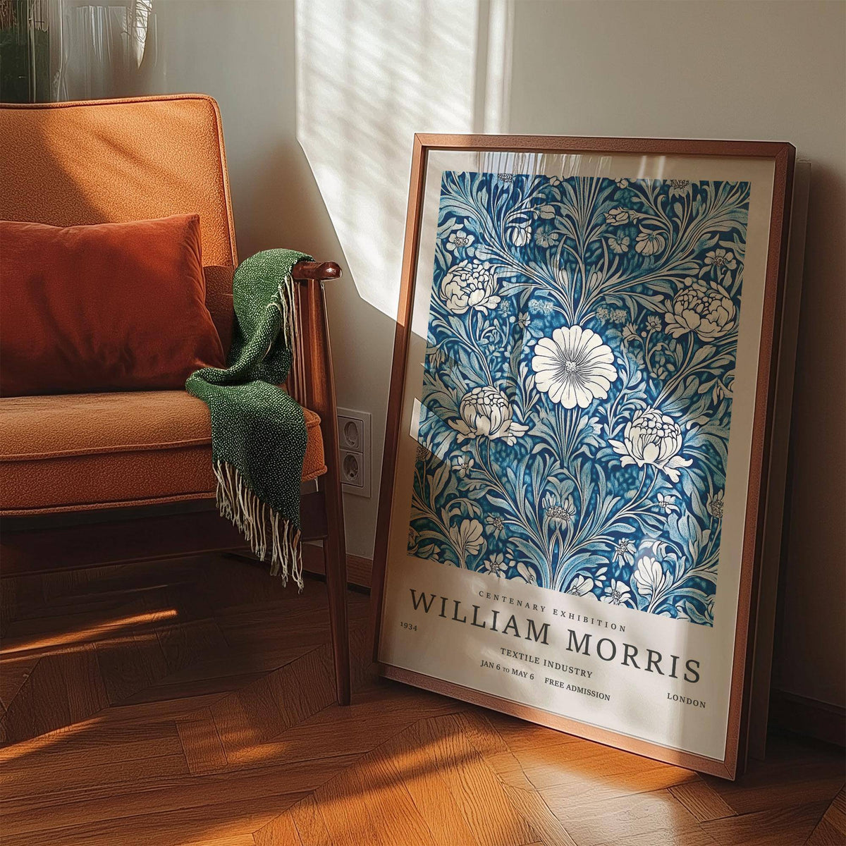 Blue botanical prints leaning on the floor with wooden frame near an orange chair and green throw