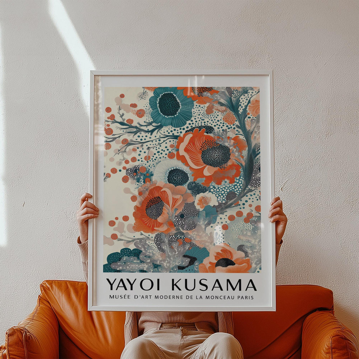 Framed print of teal and orange stylized floral blooms held against a white wall above an orange chair