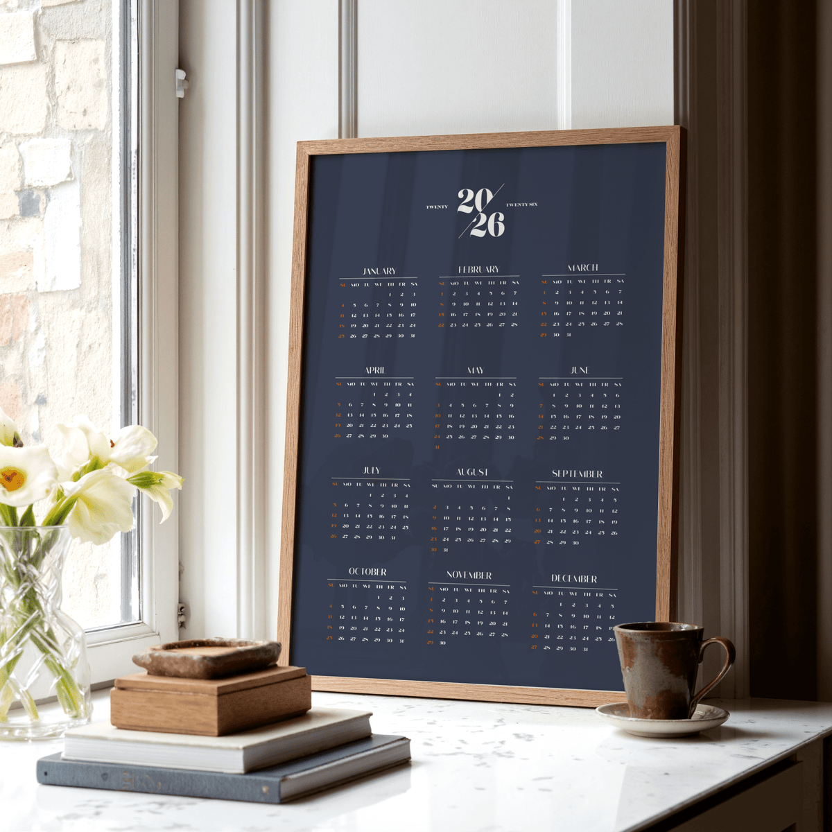 Minimalist Calendar framed print with navy background and white and orange text leaning on a window sill with flowers