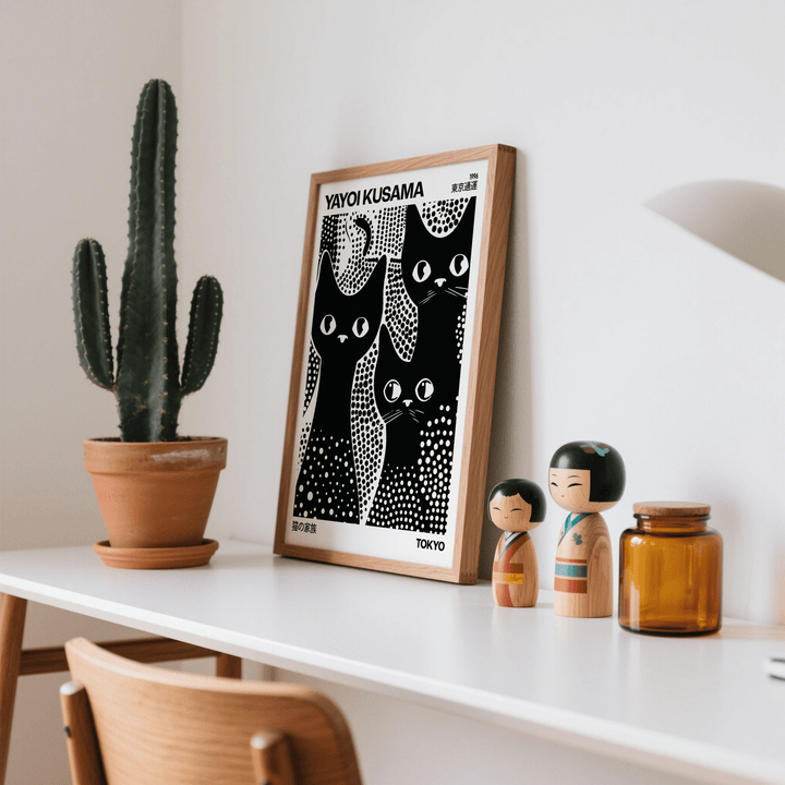 Framed Black Cats Poster in black and white leaning on a white surface next to two wooden dolls and a cactus