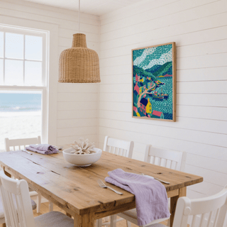 Colorful Coastal Art Print hung on a white shiplap wall above a wooden dining table with chairs and woven pendant light