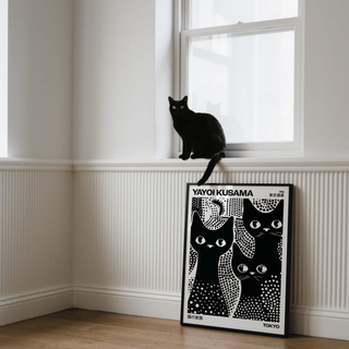 Black Cats Poster leaning on a white wall beneath a window with a real black cat on the sill