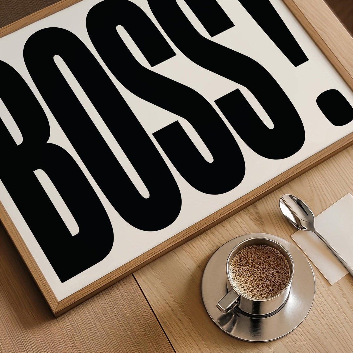 Close-up of office wall art with bold black boss typography above a coffee cup on a wooden surface