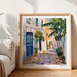 Framed Morocco wall art of a Mediterranean street scene with blue doors and lush greenery leaning on wooden floor near a sofa