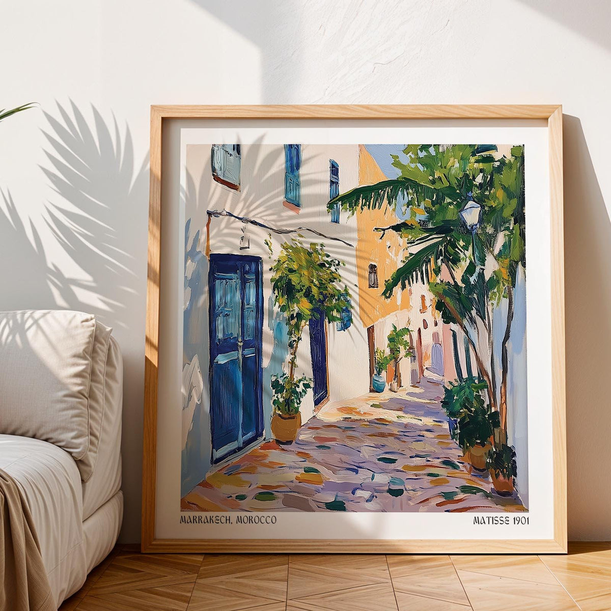 Framed Morocco wall art of a Mediterranean street scene with blue doors and lush greenery leaning on wooden floor near a sofa