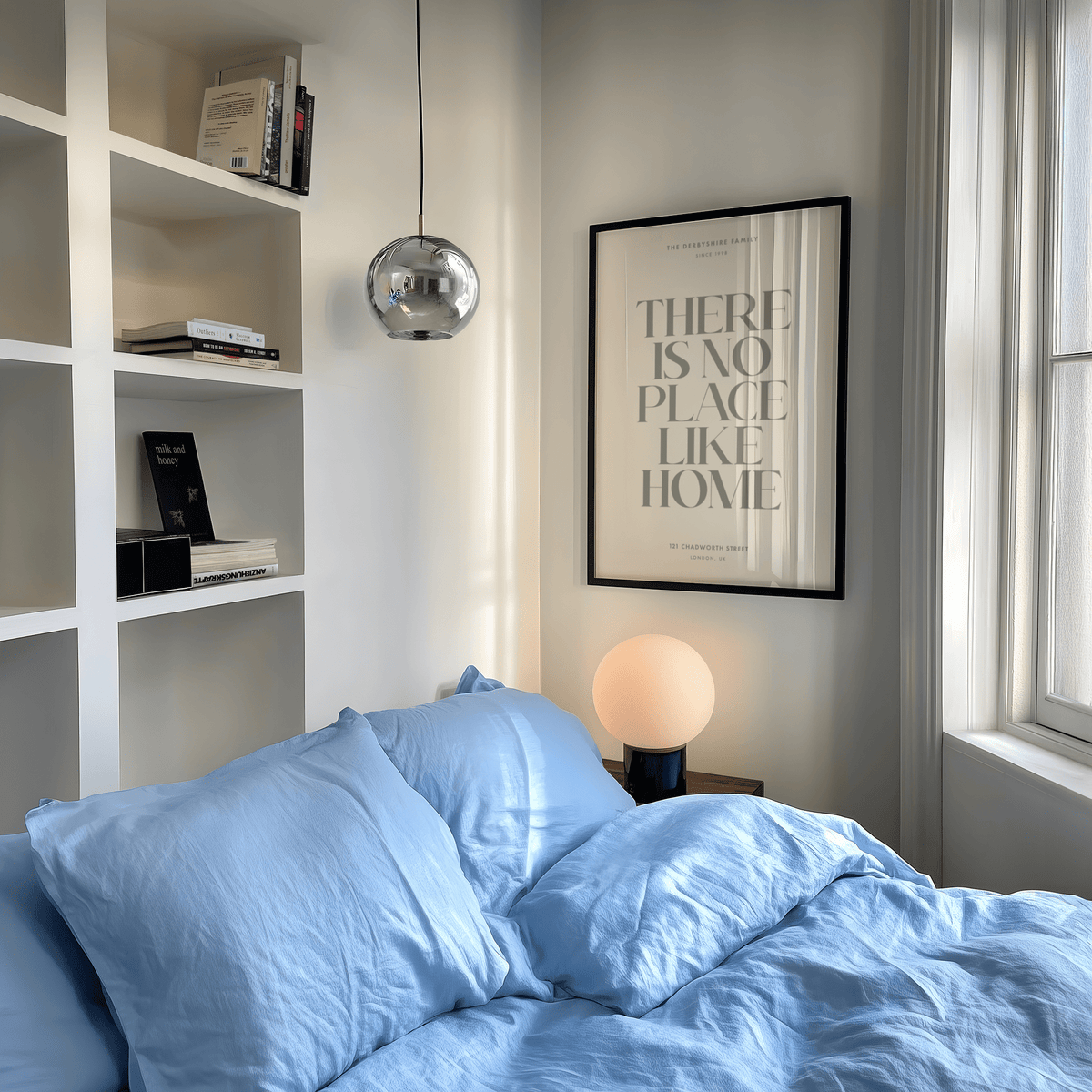 No Place Like Home print on a wall with black frame and grey text beside a bed with blue bedding and bedside lamp