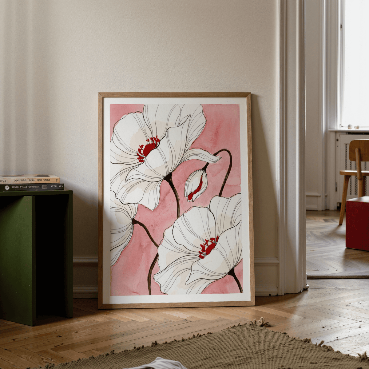 Print leaning on a wooden floor of white poppy flowers with red and pink watercolour background and a green side table with books