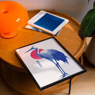 Blue Crane Art framed print in black showing a blue crane illustration with a red sun on a wooden table with an orange lamp