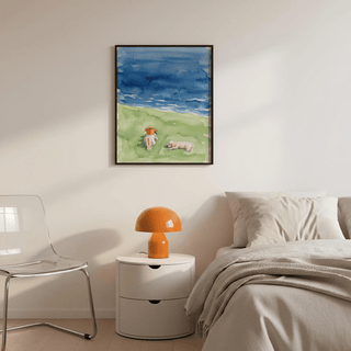 Reading By The Sea art print on wall above bedside table with orange lamp and clear chair, blue and green watercolour