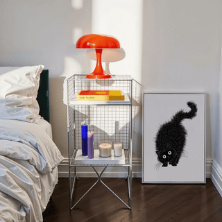 Arched Back Cat Print leaning against a wall next to a wire side table with an orange lamp in a bedroom