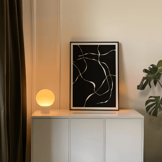 Abstract Lines art print with white abstract lines on black leaning on a white cabinet with lamp and plant