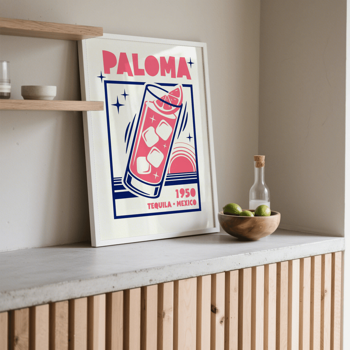 Print leaning on a shelf Paloma Cocktail Print with pink and navy mid-century style beside a wooden bowl of limes