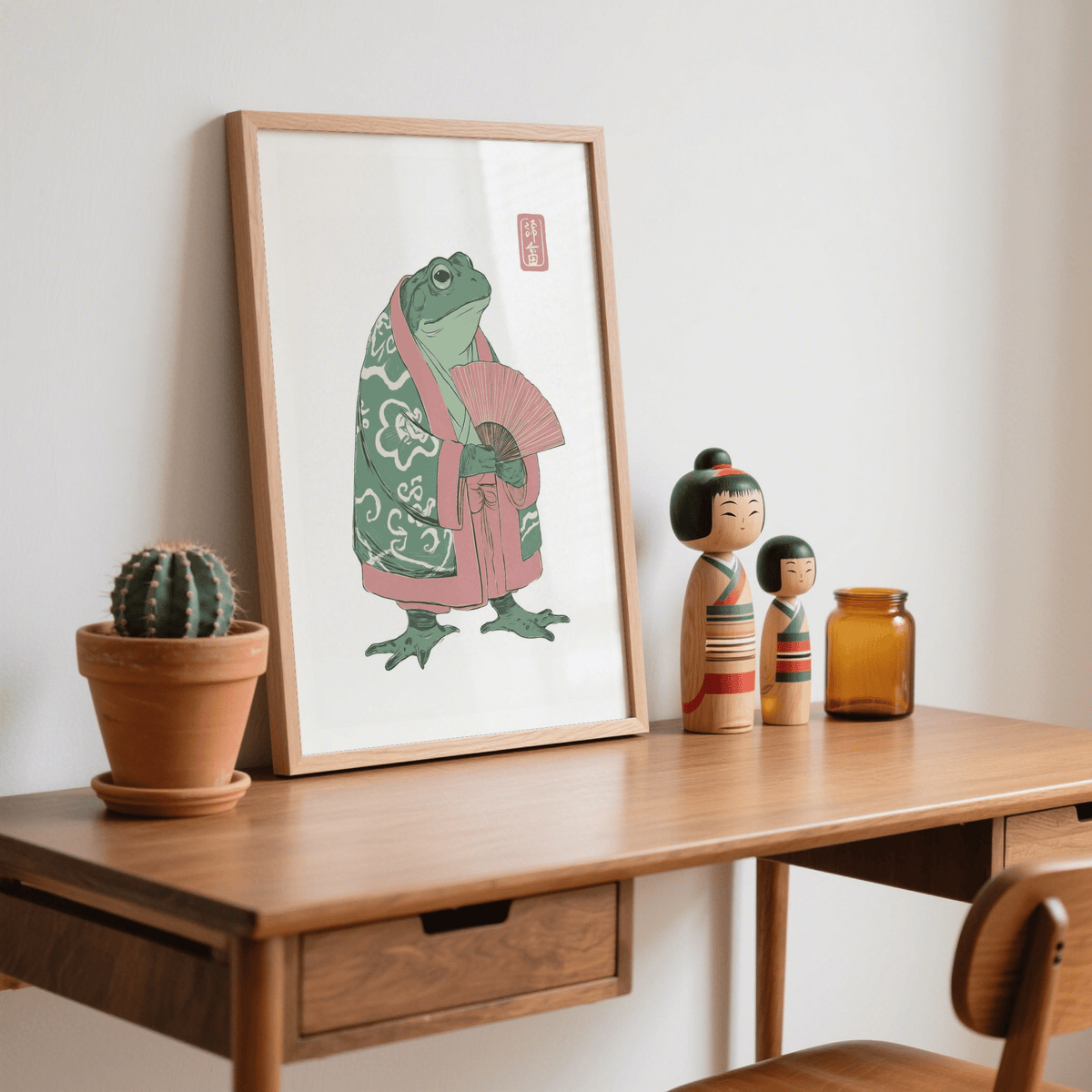 Print leaning on a wooden desk of a frog in a pink and green kimono with a fan next to a cactus and wooden dolls