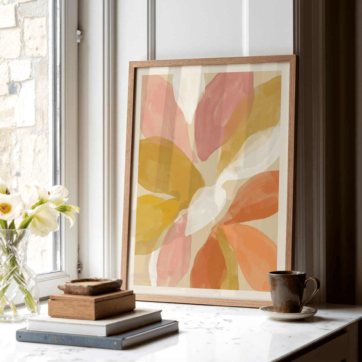 Floral Watercolor Art Print leaning on a white marble windowsill with yellow orange and pink petals next to books and a brown cup