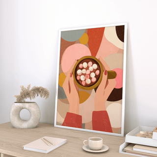Print leaning on a desk of hands holding hot chocolate with marshmallows in soft warm colours with a decorative vase nearby