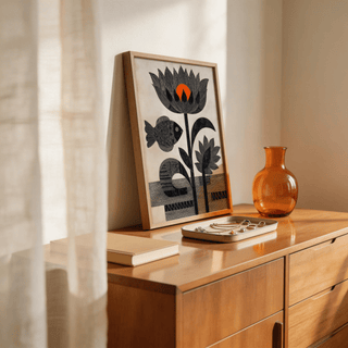Print leaning on a wooden dresser featuring black fish and flowers with an orange detail and a glass vase nearby
