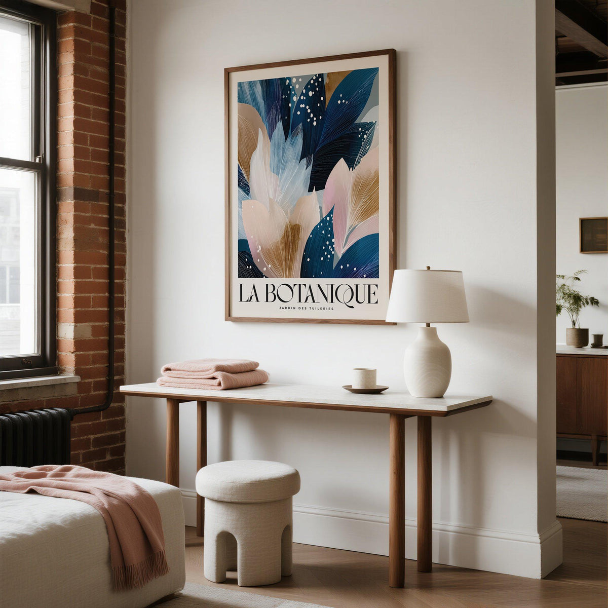 La Botanique print leaning above a wooden table with a white lamp and soft pink textiles in a bedroom