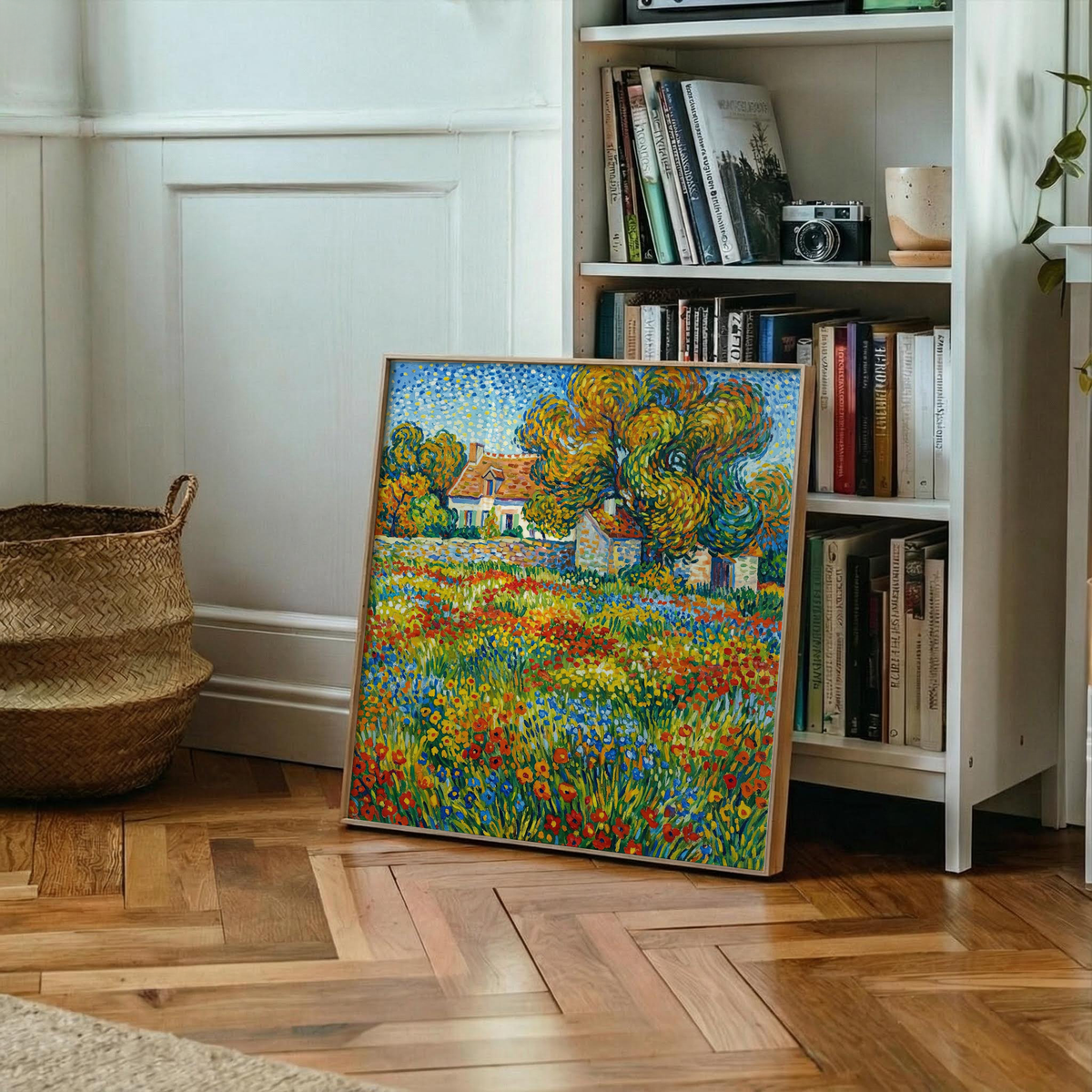 Print leaning on floor of colourful floral landscape art with houses and trees next to a bookshelf and wicker basket