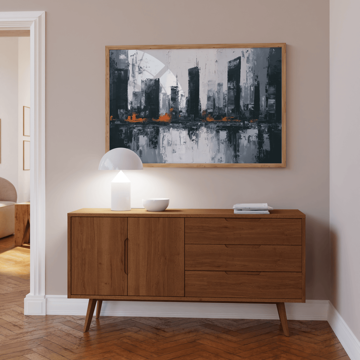 Abstract city wall art with dark grey buildings and orange highlights above wooden sideboard with lamp