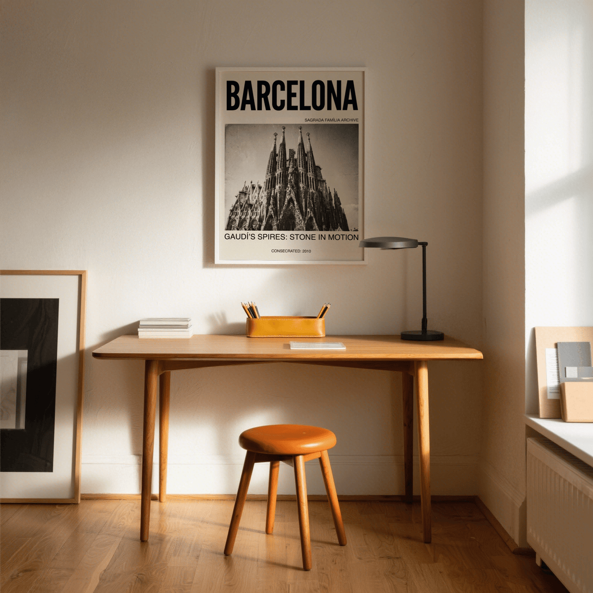 Print on a wall of Barcelona Architecture Poster showing vintage black and white Sagrada Familia spires above a wooden desk with a lamp