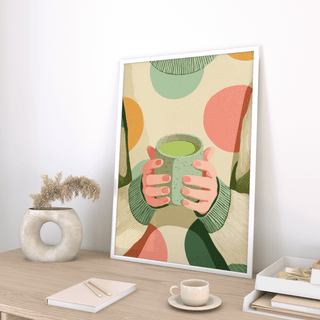 Print leaning on a wooden desk of hands holding a matcha cup with warm colours and abstract background above a small cup