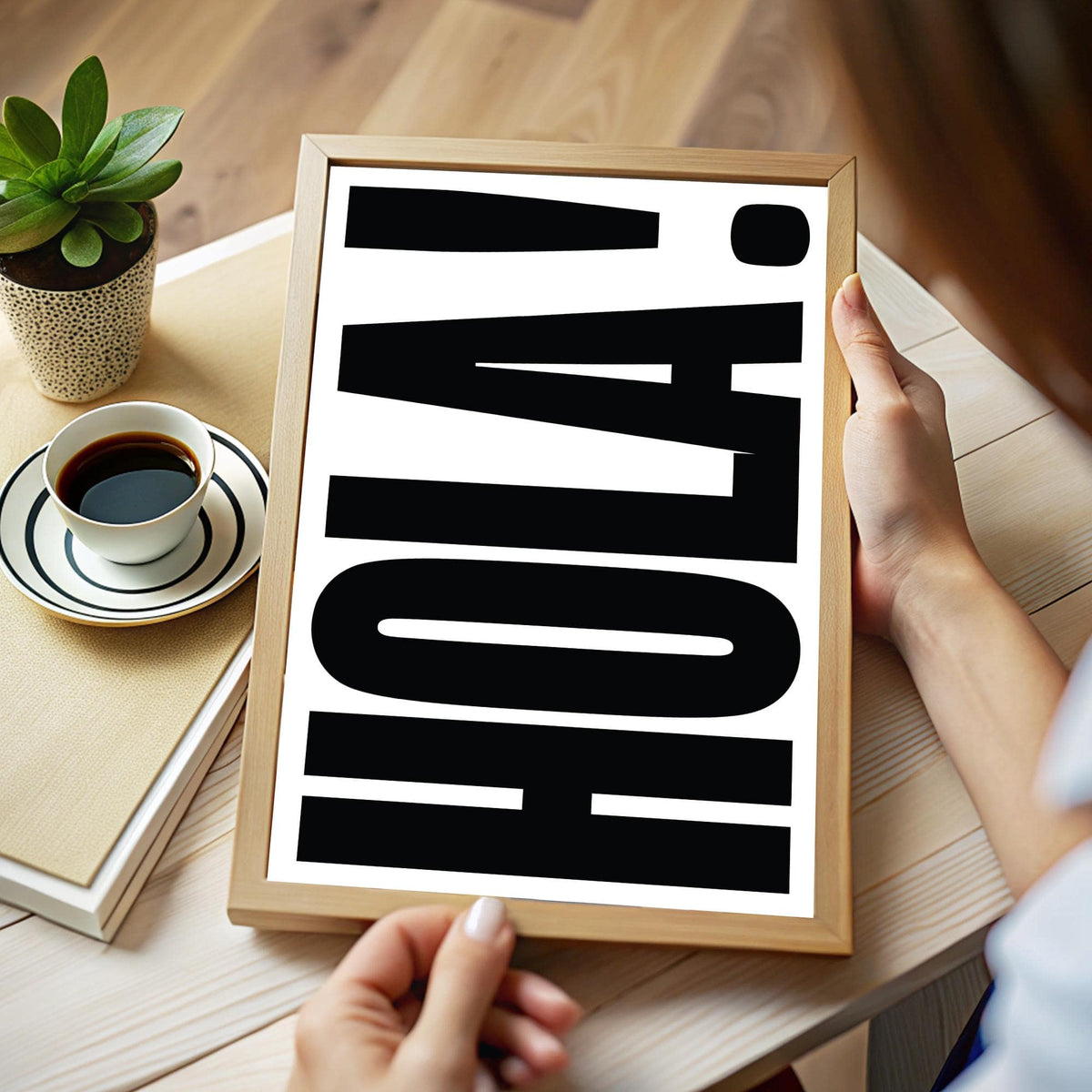 Framed close-up of HOLA typography print in black on white held above a wooden table with a coffee cup and plant