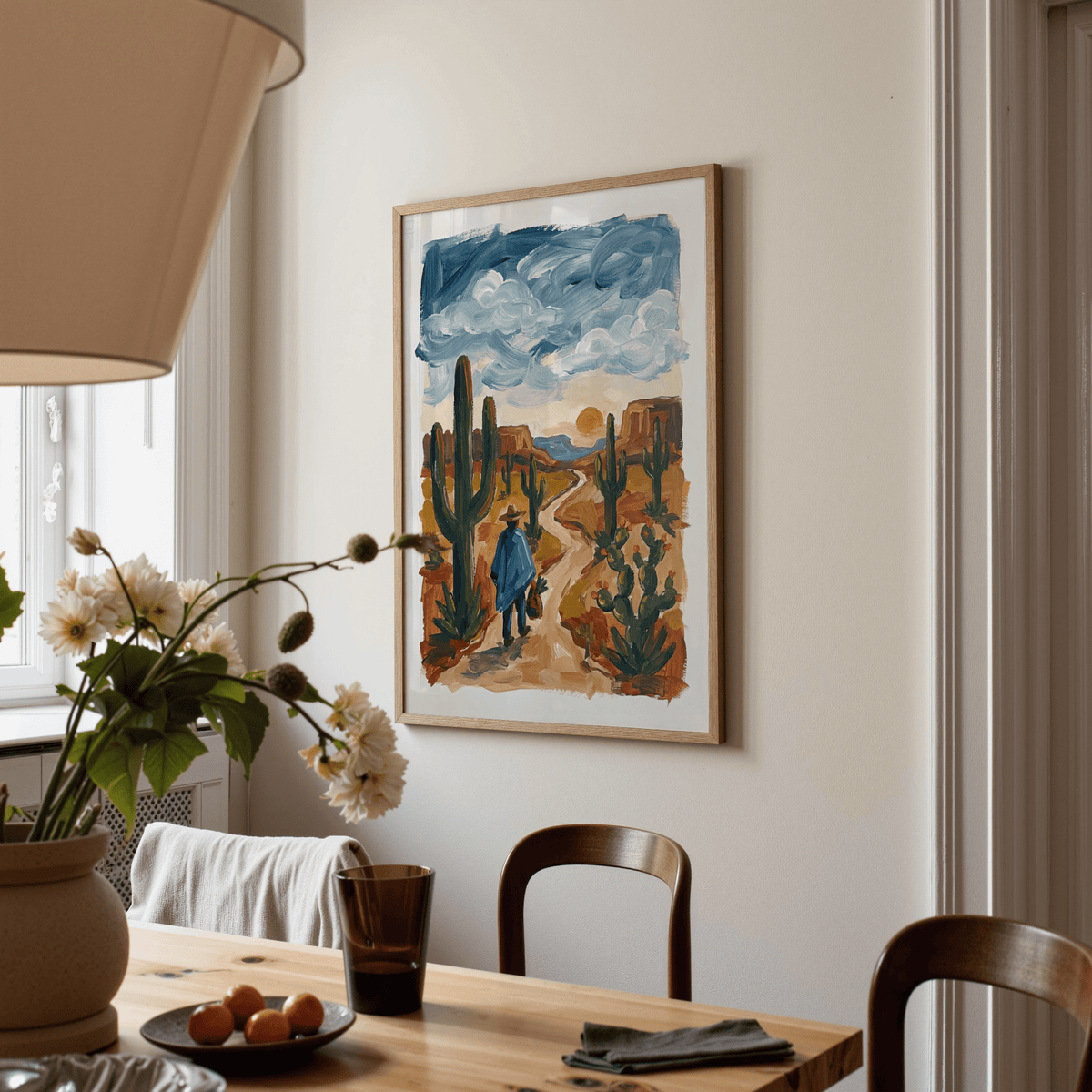 Print on a wall of a figure walking among cactuses in sunset tones above dining table with flowers