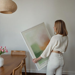 Person holding abstract sunset landscape print with soft green and orange hues near dining table with flowers