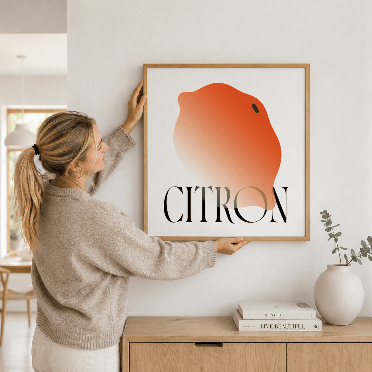 Framed Abstract Lemon Art Print with orange gradient and black text held by a woman above a wooden sideboard