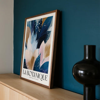 Framed La Botanique Print with abstract floral leaves in blues and browns leaning on a shelf with a black vase