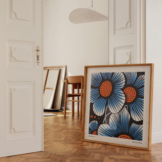 Print leaning on floor with blue flowers and orange centres near white double doors and wooden chair