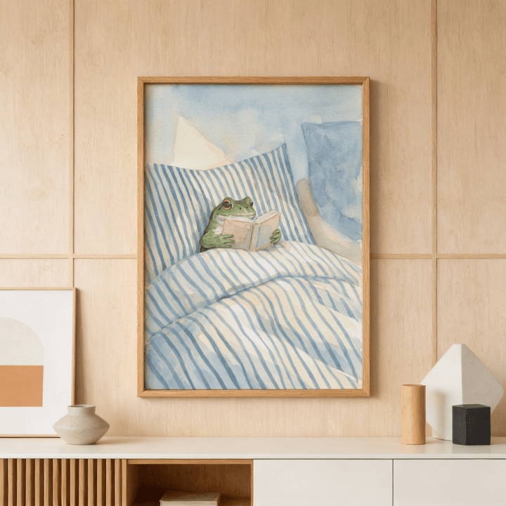 Frog Reading In Bed print with blue striped bedding in a wooden framed print above a sideboard with vase