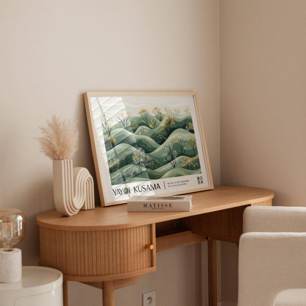 Print leaning on a wooden desk showing minimalist hills with blossoming trees in green and yellow hues with a book nearby