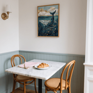 Whale wall art of two whales underwater in blue and gold hangs above a dining table with wooden chairs